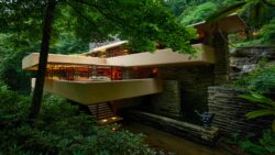 The southeast elevation of the house at Fallingwater photographed from the bridge. The house lights are on and cast a warm glow through the large windows. The house is surrounded by green foliage of the trees and rhododendrons and the water of Bear Run flows below the house before reaching the falls on the southwest side of the house.