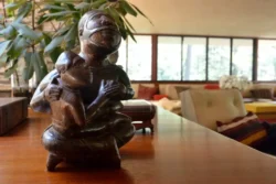 A brown stylized sculpture of a mother holding a child sits on top of a wooden desk in the Fallingwater living room. Rhododendron leaves and windows are in the background.