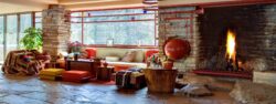 A fire roars atop the stone hearth of the living room fireplace at Fallingwater. A large red cauldron is nestled into the stone masonry next to the fireplace. A built-in sofa lines the wall below large windows, covered in various textiles.
