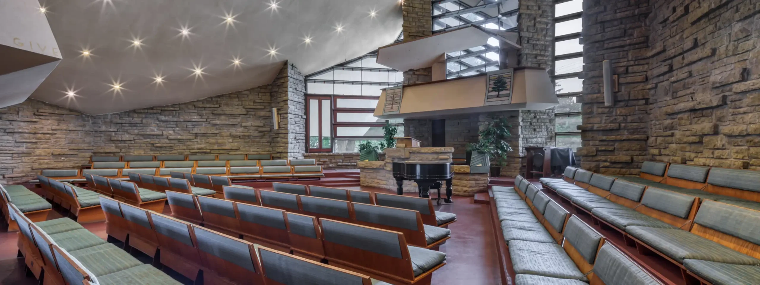 Pews extend toward a pulpit in front of large windows. Stone walls support a cantilevered roof that extends beyond the pulpit and windows.
