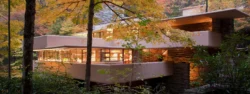 The beige terraces of a modern house (Fallingwater) extend over a stream. The surrounding forest has turned to a Fall palette of yellows, oranges and reds.