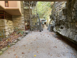 A man builds scaffolding along the northeastern wall of Fallingwater. There is a large boulder to his right.